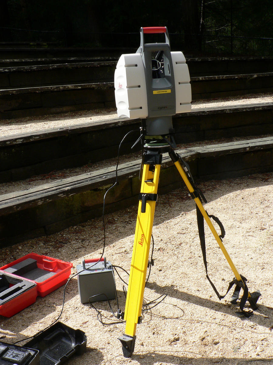 The Leica terrestrial LIDAR scanner is set up on a tripod. There is some kind of scaffolding behind it, a small red box of equipment to the left and it is sitting on a flat sandy surface.