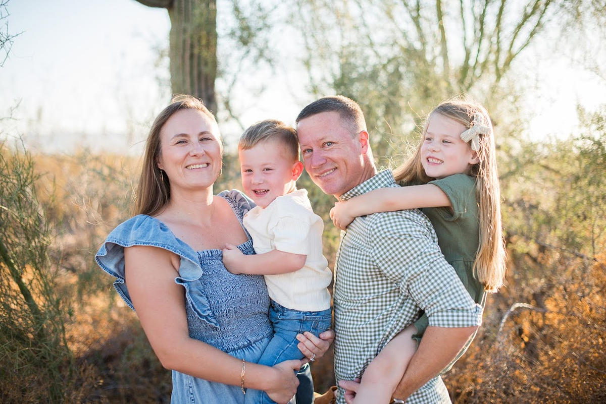 A photo of Phil Orstrom and his family in the Arizona desert. His wife is on the left hold his son to her right. Phil is to the right of his son facing his wife, and his daughter is riding piggy back.  They are all smiling. 